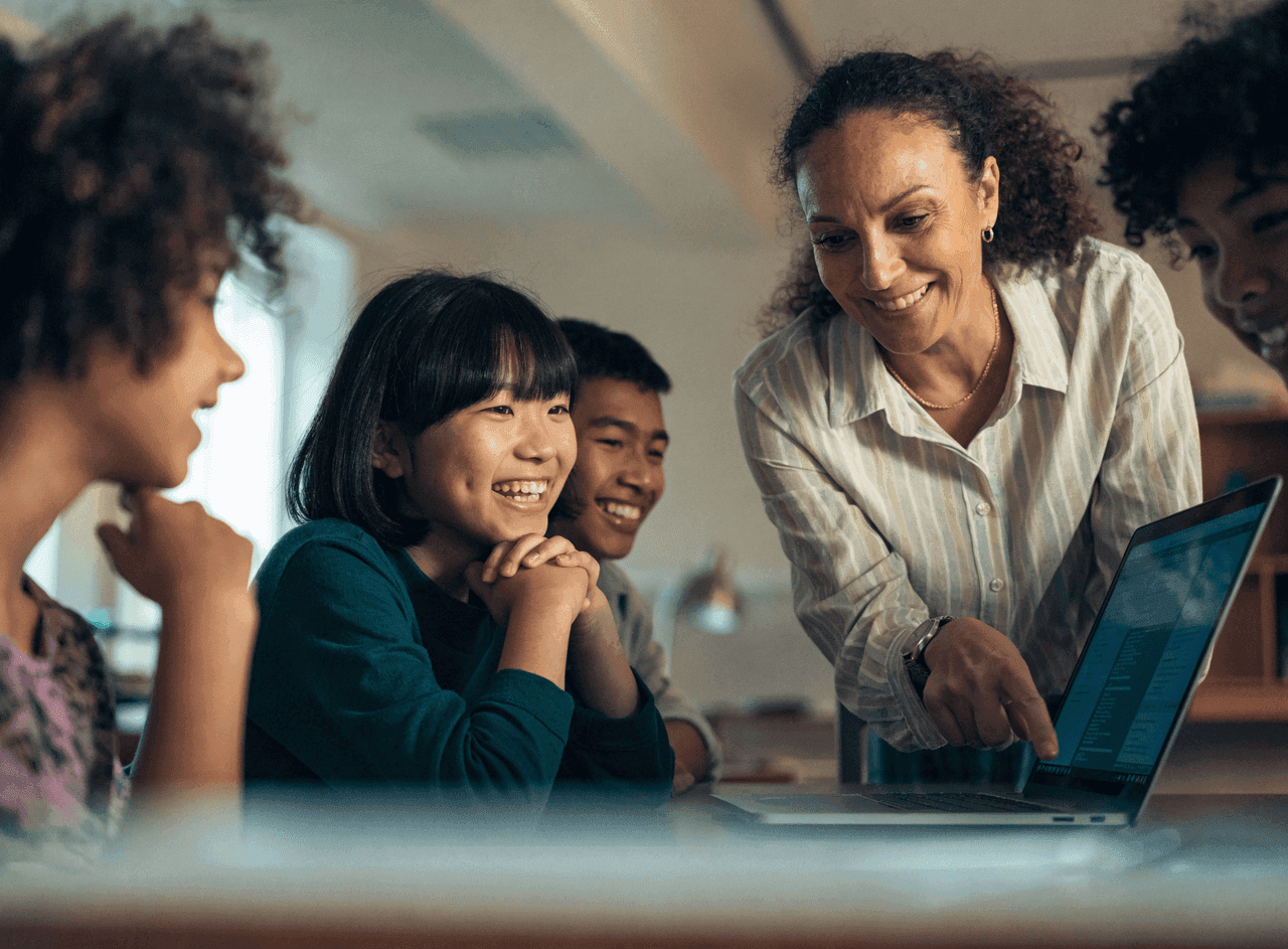 A teacher smiling and guiding students around a laptop in a bright modern classroom